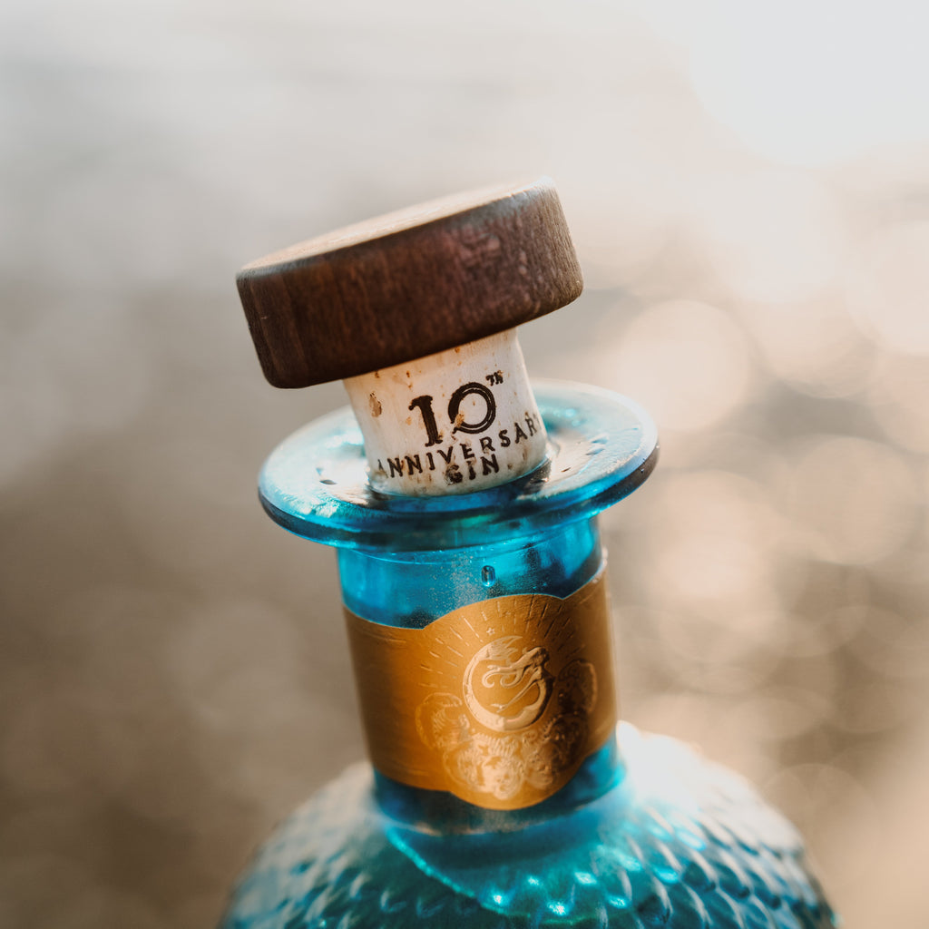 Close-up of a blue bottle with a cork labeled '10th Anniversary Gin' against a blurred background.