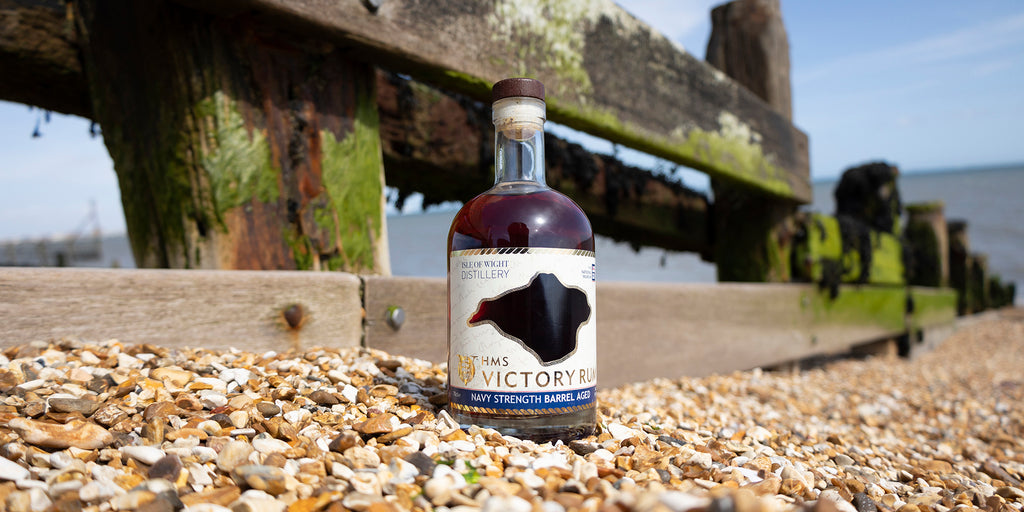 HMS Victory Strength Rum from the Isle of Wight Distillery, on a pebble beach with the ocean and seaweed covered groynes in view. 
